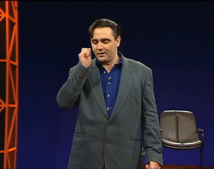 Tony Slattery performing on stage in a suit with a chair in the background. Tony Slattery performing on stage in a suit with a chair in the background.