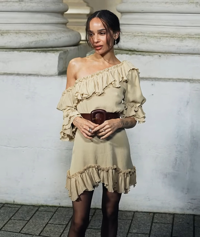 Zoë Kravitz in a beige dress, brown belt, and black stockings posing for a photo. Zoë Kravitz in a beige dress, brown belt, and black stockings posing for a photo.