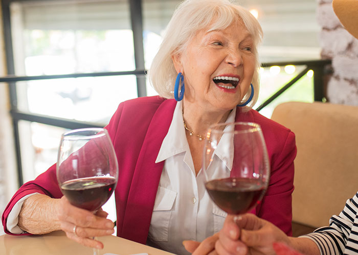 Elderly woman in a pink blazer laughing and holding a glass of red wine at a social gathering. Elderly woman in a pink blazer laughing and holding a glass of red wine at a social gathering.