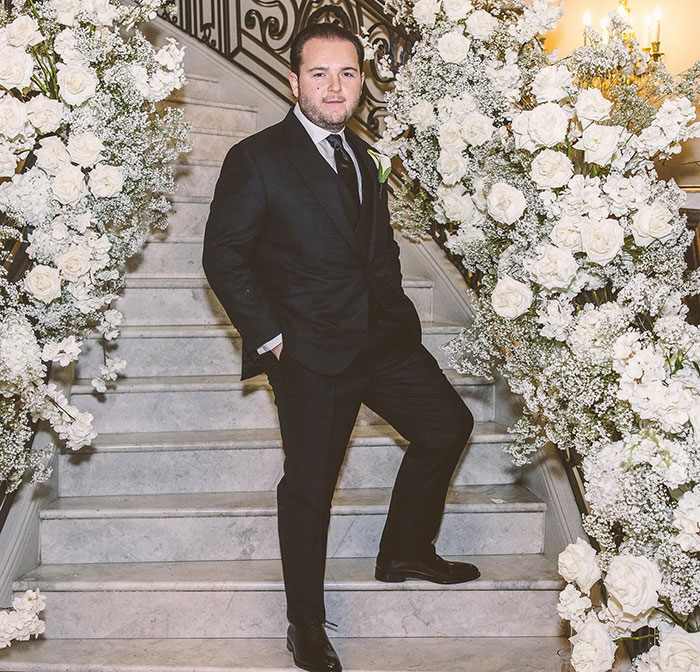 Man in a suit standing on flower-decorated stairs, related to School of Rock child stars reunion. Man in a suit standing on flower-decorated stairs, related to School of Rock child stars reunion.