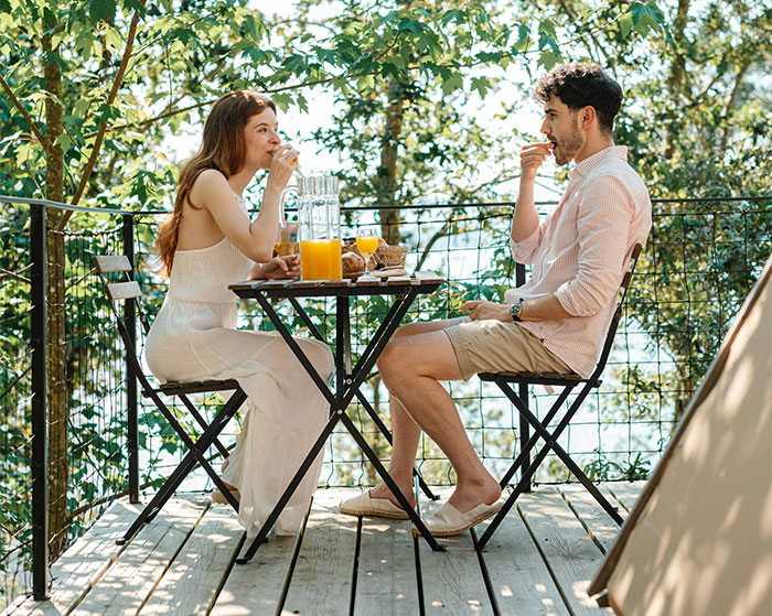 A couple dining outdoors, enjoying a breakfast with juice, surrounded by lush trees on a wooden deck. A couple dining outdoors, enjoying a breakfast with juice, surrounded by lush trees on a wooden deck.