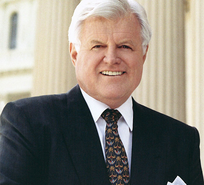 A person in a formal suit and tie, standing outside a building, reflecting celebrity secrets and scandals.