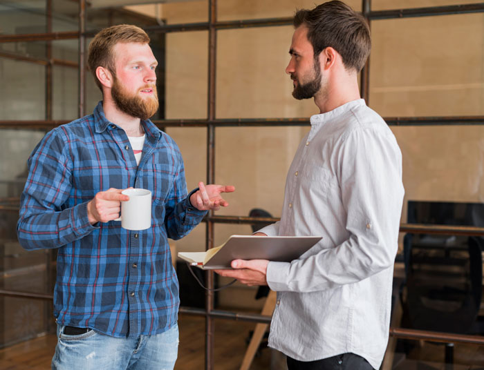 Two men in conversation, one holding a laptop and the other a mug, discussing wedding dress shopping. Two men in conversation, one holding a laptop and the other a mug, discussing wedding dress shopping.