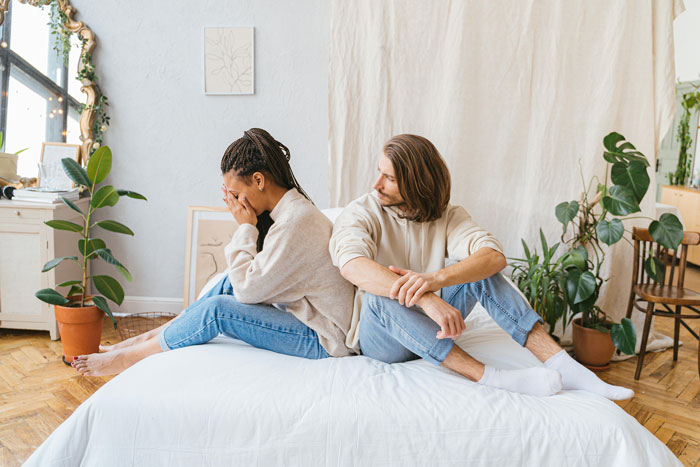 A couple sitting back-to-back on a bed, looking upset during wedding dress shopping day. A couple sitting back-to-back on a bed, looking upset during wedding dress shopping day.