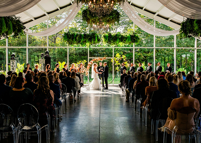 Newlyweds at an indoor wedding ceremony with guests seated, featuring elegant decor and lush greenery. Newlyweds at an indoor wedding ceremony with guests seated, featuring elegant decor and lush greenery.