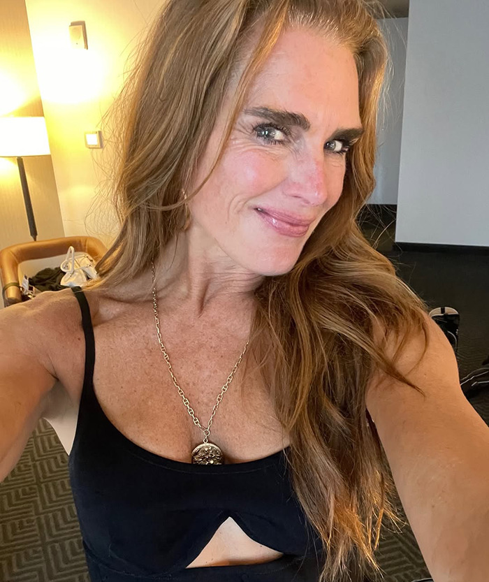 Brooke Shields with long hair and necklace, wearing a black top, smiling in a casual indoor setting. Brooke Shields with long hair and necklace, wearing a black top, smiling in a casual indoor setting.