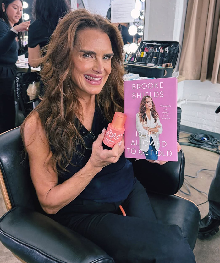 Brooke Shields holding her book and a skincare product, smiling in a dressing room setting. Brooke Shields holding her book and a skincare product, smiling in a dressing room setting.