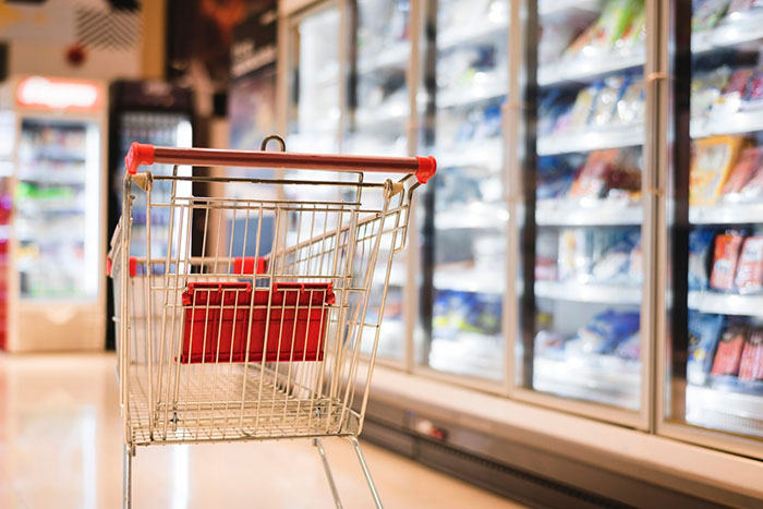 An empty shopping cart in a grocery store aisle with freezer section on the right. An empty shopping cart in a grocery store aisle with freezer section on the right.