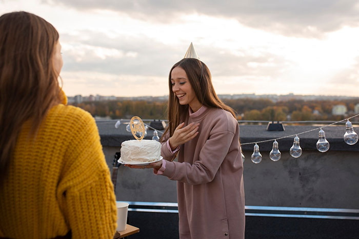 Woman holding a cake while smiling at a rooftop gathering, wearing a party hat, with a background of lights and trees. Woman holding a cake while smiling at a rooftop gathering, wearing a party hat, with a background of lights and trees.