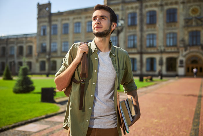 Male student outside a university building, carrying books, related to college funding challenges. Male student outside a university building, carrying books, related to college funding challenges.