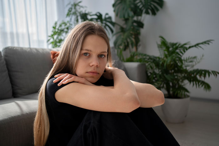 Teen expressing annoyance while sitting alone on a couch, surrounded by plants in a sunlit room. Teen expressing annoyance while sitting alone on a couch, surrounded by plants in a sunlit room.