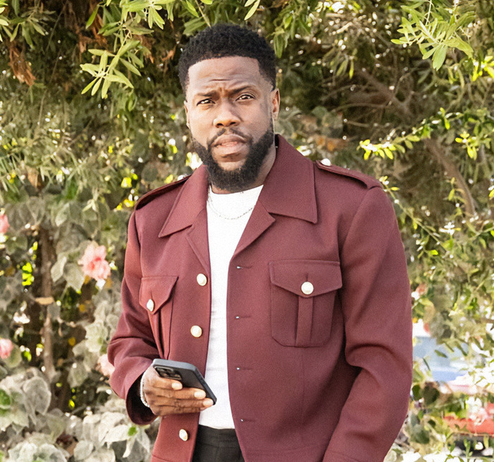 Kevin Hart outdoors holding phone, wearing maroon jacket, amid greenery. Kevin Hart outdoors holding phone, wearing maroon jacket, amid greenery.
