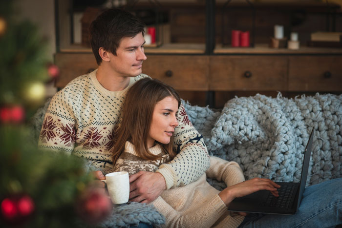 A couple on a sofa, wearing winter sweaters, with a laptop and mug, depicting family New Year's Eve drama. A couple on a sofa, wearing winter sweaters, with a laptop and mug, depicting family New Year's Eve drama.