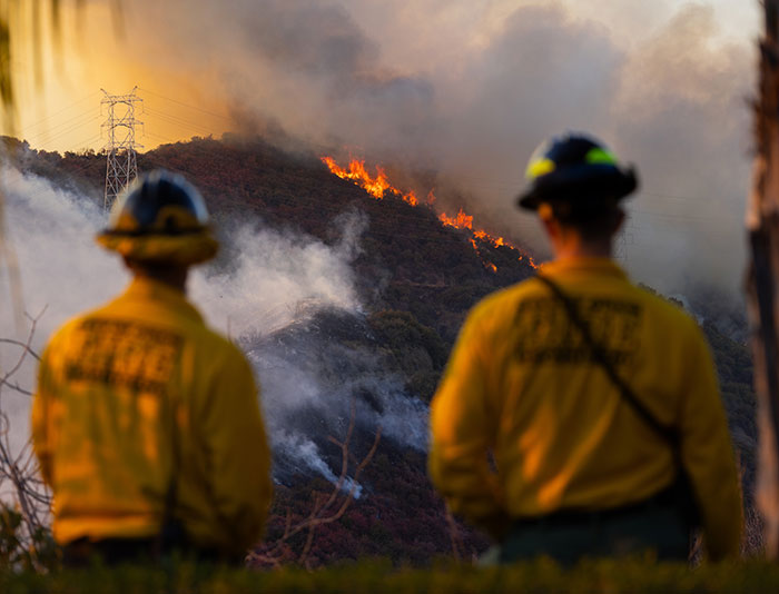 Private firefighters observe wildfire approaching a hillside, smoke and flames visible. Private firefighters observe wildfire approaching a hillside, smoke and flames visible.