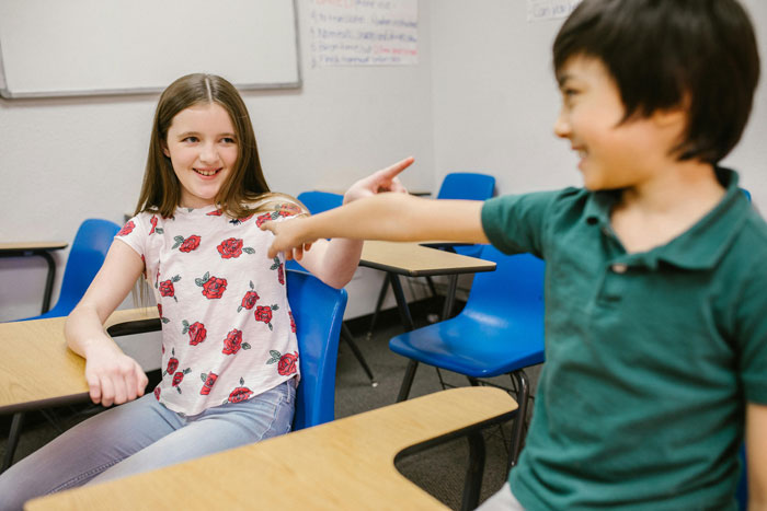 Two children pointing at each other, seated in a classroom with blue chairs, laughter in the air. Two children pointing at each other, seated in a classroom with blue chairs, laughter in the air.
