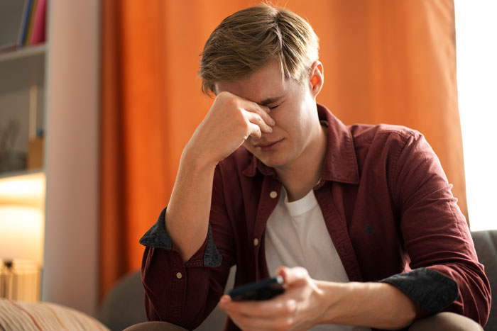 Teen holding phone, looking upset, wearing a maroon shirt, sitting in a room with orange curtains. Teen holding phone, looking upset, wearing a maroon shirt, sitting in a room with orange curtains.