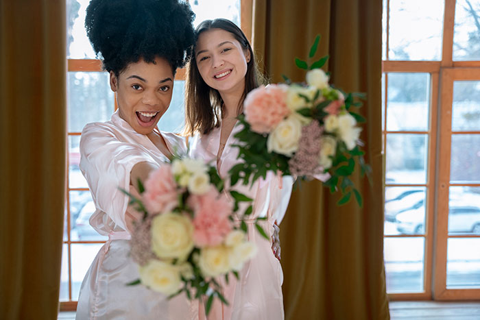 Two women in robes joyfully holding floral bouquets indoors near large windows. Two women in robes joyfully holding floral bouquets indoors near large windows.