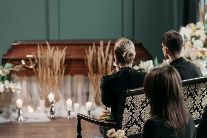 People in black outfits at a somber gathering, facing a casket surrounded by candlelit silverware. People in black outfits at a somber gathering, facing a casket surrounded by candlelit silverware.