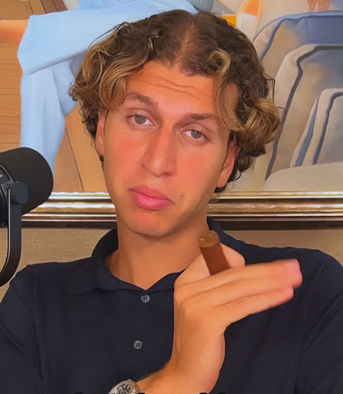 Person holding a cigar, wearing a dark polo, sits near a microphone. Parodying billionaires online. Person holding a cigar, wearing a dark polo, sits near a microphone. Parodying billionaires online.