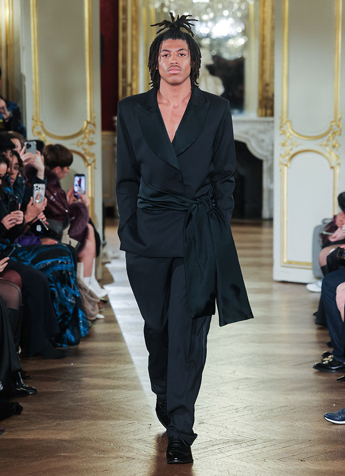 Young male model making runway debut at Paris Fashion Week in a black suit, walking confidently on a wooden floor. Young male model making runway debut at Paris Fashion Week in a black suit, walking confidently on a wooden floor.