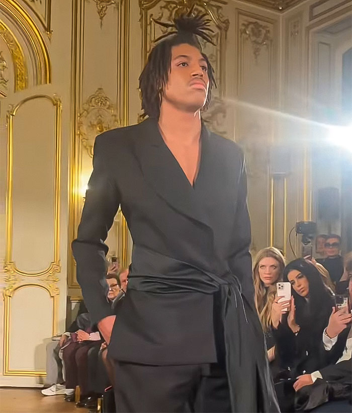 Model walks runway at Paris Fashion Week in sleek black suit, with audience in the background. Model walks runway at Paris Fashion Week in sleek black suit, with audience in the background.