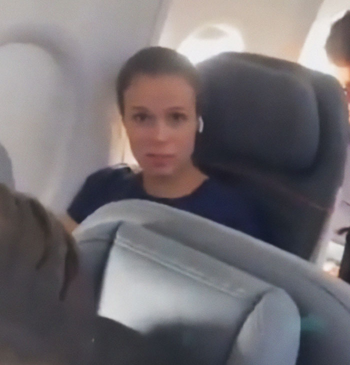 Woman in airplane seat, involved in seat swap dispute with crying child, wearing navy shirt and earbuds. Woman in airplane seat, involved in seat swap dispute with crying child, wearing navy shirt and earbuds.
