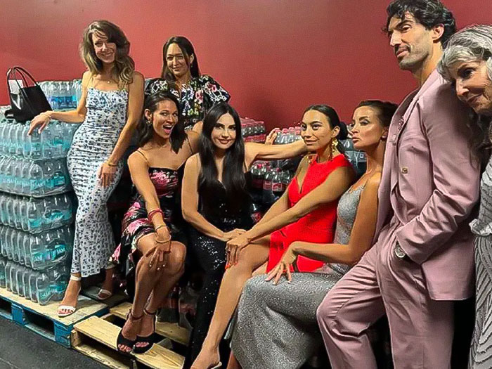 Group posing in a basement setting, Justin Baldoni in a pink suit beside water bottles, surrounded by smiling people. Group posing in a basement setting, Justin Baldoni in a pink suit beside water bottles, surrounded by smiling people.