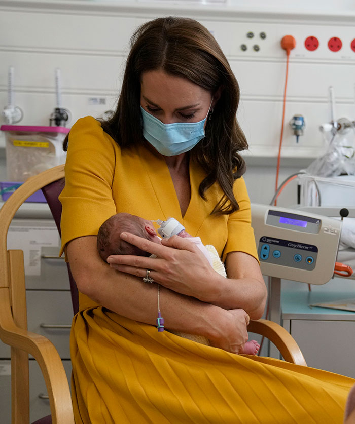 A woman in a yellow dress, wearing a mask, holds a baby in a hospital setting, engagement ring visible. A woman in a yellow dress, wearing a mask, holds a baby in a hospital setting, engagement ring visible.