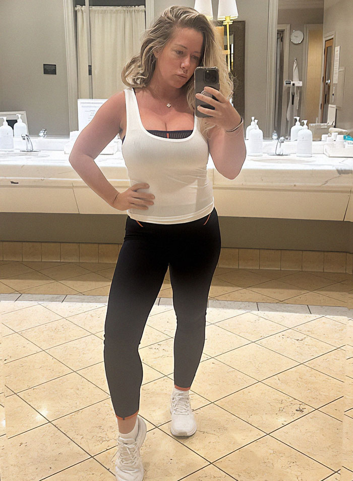 A woman takes a selfie in a bathroom wearing a white tank top and black leggings, addressing weight gain criticism. A woman takes a selfie in a bathroom wearing a white tank top and black leggings, addressing weight gain criticism.