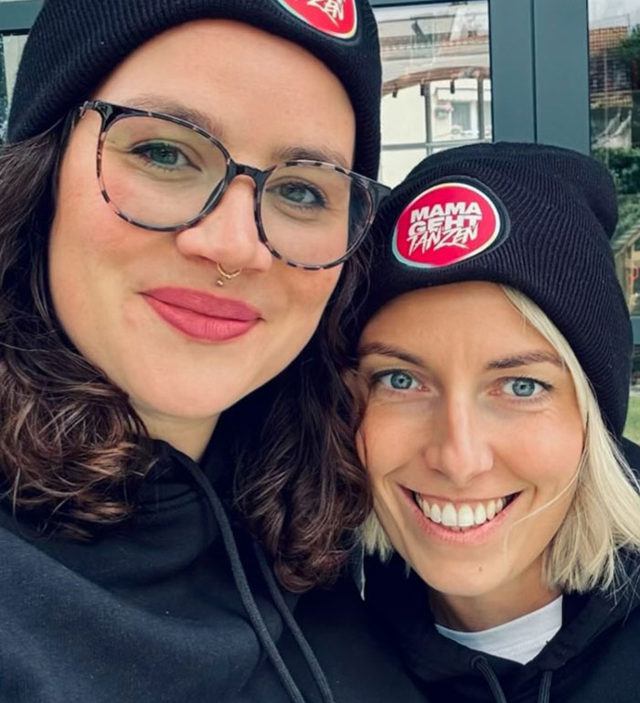 Two moms wearing matching beanies with the logo "Mama geht tanzen," smiling and redefining post-baby partying. Two moms wearing matching beanies with the logo "Mama geht tanzen," smiling and redefining post-baby partying.