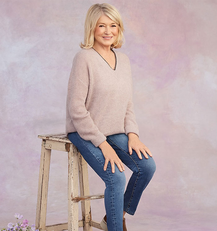 An individual in a light sweater and jeans sits on a rustic stool against a soft, pastel backdrop. An individual in a light sweater and jeans sits on a rustic stool against a soft, pastel backdrop.