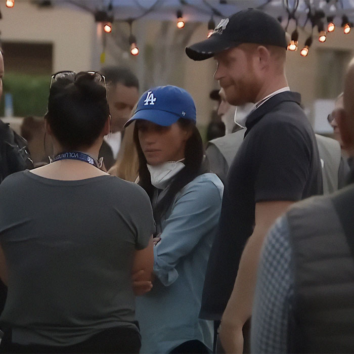 A woman in a blue cap stands with a man in a black cap, engaged in a conversation; Meghan Markle, Prince Harry related scene. A woman in a blue cap stands with a man in a black cap, engaged in a conversation; Meghan Markle, Prince Harry related scene.