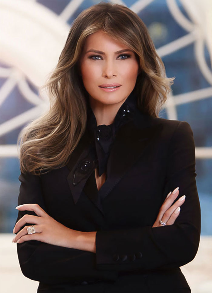 First Lady portrait with Melania Trump in a black blazer, posing with arms crossed against a blurred background. First Lady portrait with Melania Trump in a black blazer, posing with arms crossed against a blurred background.