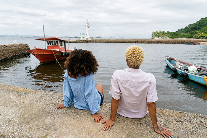 A man and woman sitting by the waterfront, boats in view, symbolizing a relationship and proposal moment in a crowd. A man and woman sitting by the waterfront, boats in view, symbolizing a relationship and proposal moment in a crowd.