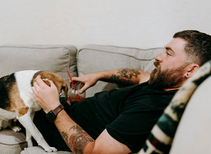 Man relaxing on a couch with a drink, petting a dog, showcasing entitled behavior. Man relaxing on a couch with a drink, petting a dog, showcasing entitled behavior.
