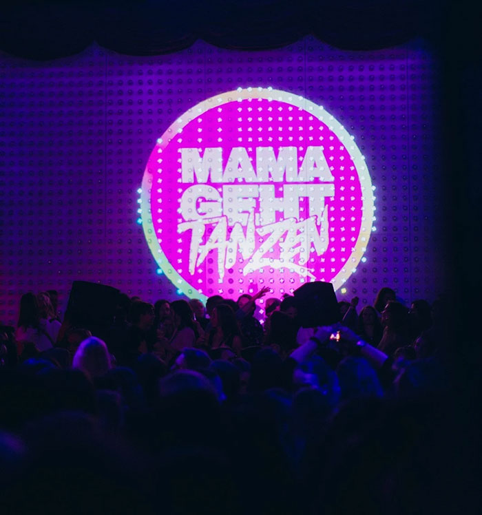 Nightclub scene with "Mama Geht Tanzen" projected on wall, redefining partying post-baby for moms. Nightclub scene with "Mama Geht Tanzen" projected on wall, redefining partying post-baby for moms.