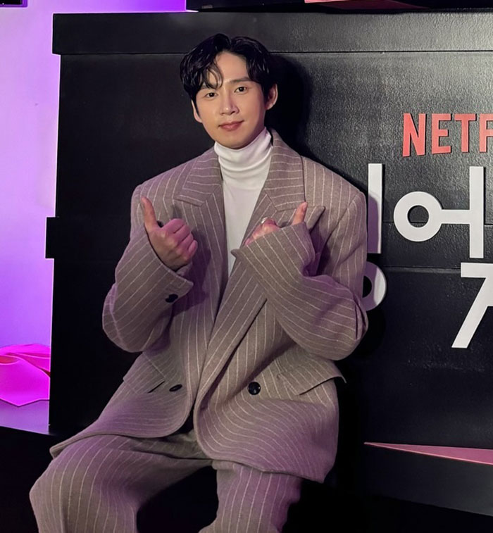 Squid Game actor in pinstripe suit, giving thumbs up at Netflix event backdrop. Squid Game actor in pinstripe suit, giving thumbs up at Netflix event backdrop.