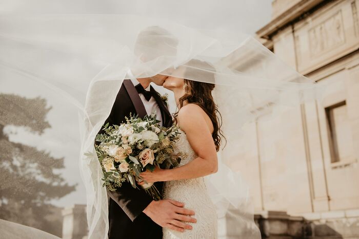 Couple in wedding attire kissing under a veil, holding a bouquet, on a sunny day.