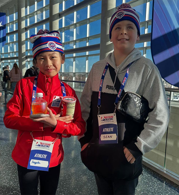 Adorable child ice skating duo wearing matching hats and badges, smiling indoors. Adorable child ice skating duo wearing matching hats and badges, smiling indoors.