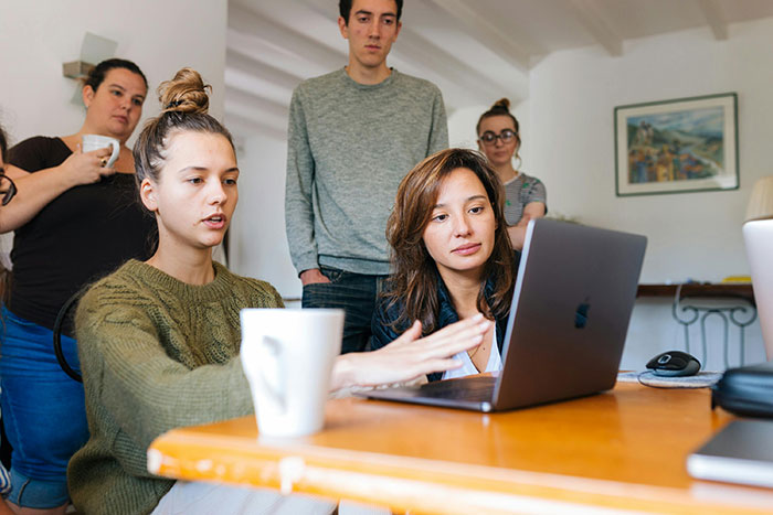 Group discussion around a laptop, two women focused, others watching, in a casual work setting. Group discussion around a laptop, two women focused, others watching, in a casual work setting.