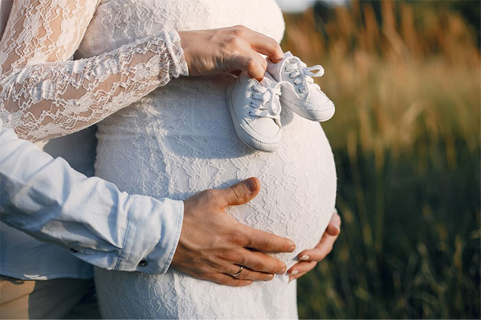 Pregnant woman in lace dress holding baby shoes, hands supporting her belly, outdoors. Pregnant woman in lace dress holding baby shoes, hands supporting her belly, outdoors.