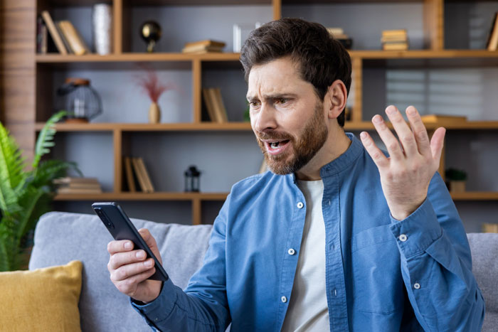 Man in blue shirt looking at phone with frustration, sitting on a sofa. Man in blue shirt looking at phone with frustration, sitting on a sofa.