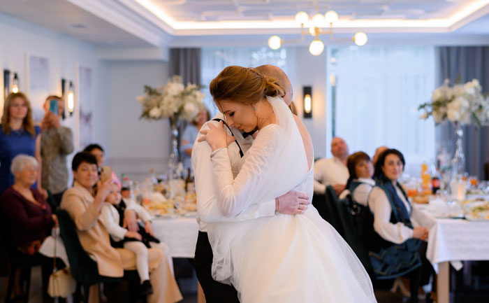 Bride embraces a guest during a wedding reception, with seated attendees and floral decorations in the background. Bride embraces a guest during a wedding reception, with seated attendees and floral decorations in the background.