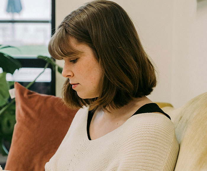 A woman with shoulder-length hair, wearing a white sweater, sits contemplatively on a couch. A woman with shoulder-length hair, wearing a white sweater, sits contemplatively on a couch.