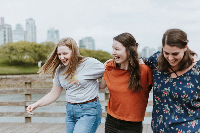Three friends laughing and walking together on a wooden path with a city skyline in the background. Three friends laughing and walking together on a wooden path with a city skyline in the background.
