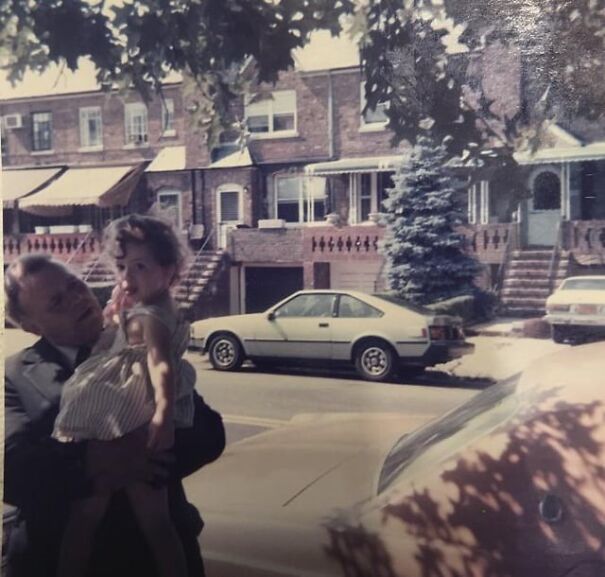 A nostalgic retro scene with a person holding a child in front of classic 80s cars and brick houses.