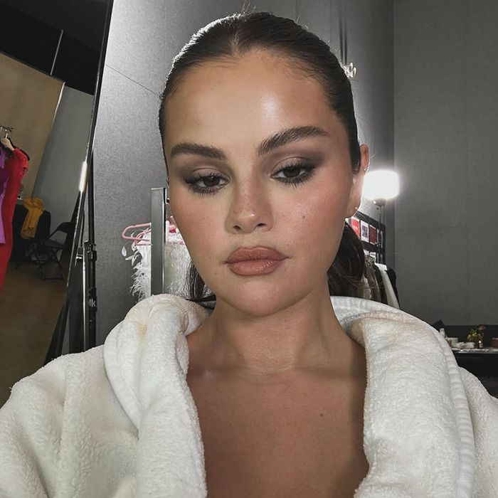 Selena Gomez in a white robe with smoky eye makeup, looking reflective under bright lights. Selena Gomez in a white robe with smoky eye makeup, looking reflective under bright lights.