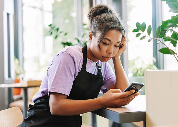 A staff member in a cafe, wearing a purple shirt and apron, looks at a phone. A staff member in a cafe, wearing a purple shirt and apron, looks at a phone.