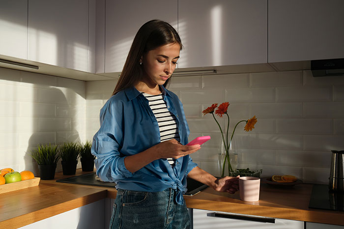 Young woman in kitchen using smartphone, holding a pink mug, with sunlight on her face. Young woman in kitchen using smartphone, holding a pink mug, with sunlight on her face.