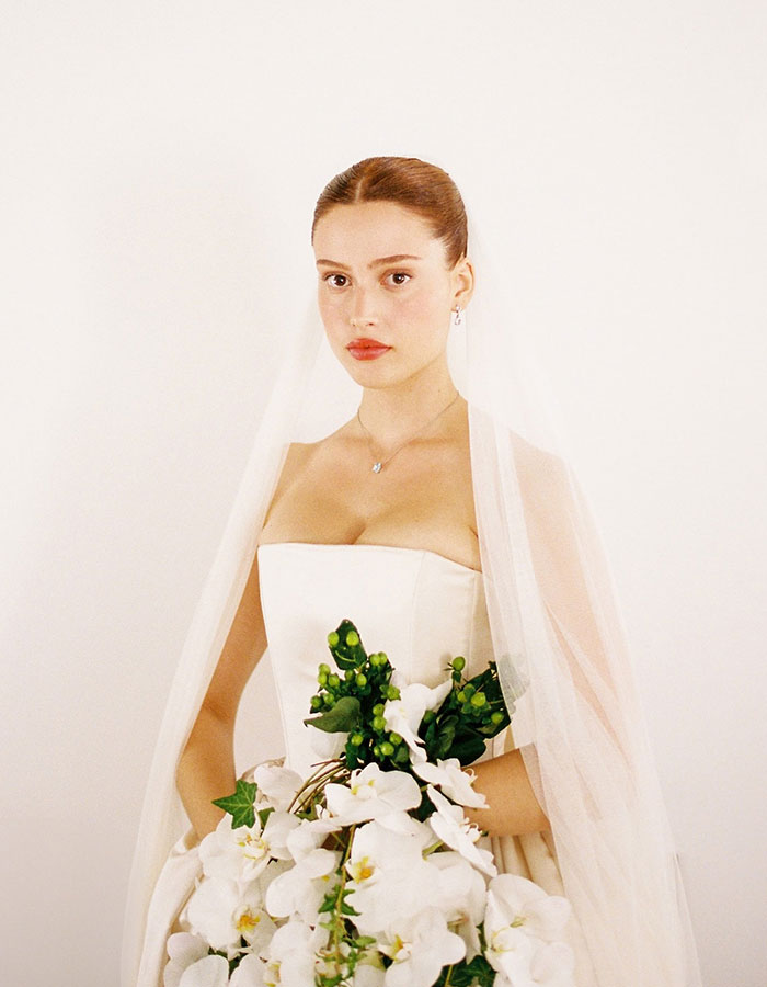 Woman in dramatic wedding dress and veil, holding white flower bouquet, standing against a plain background. Woman in dramatic wedding dress and veil, holding white flower bouquet, standing against a plain background.